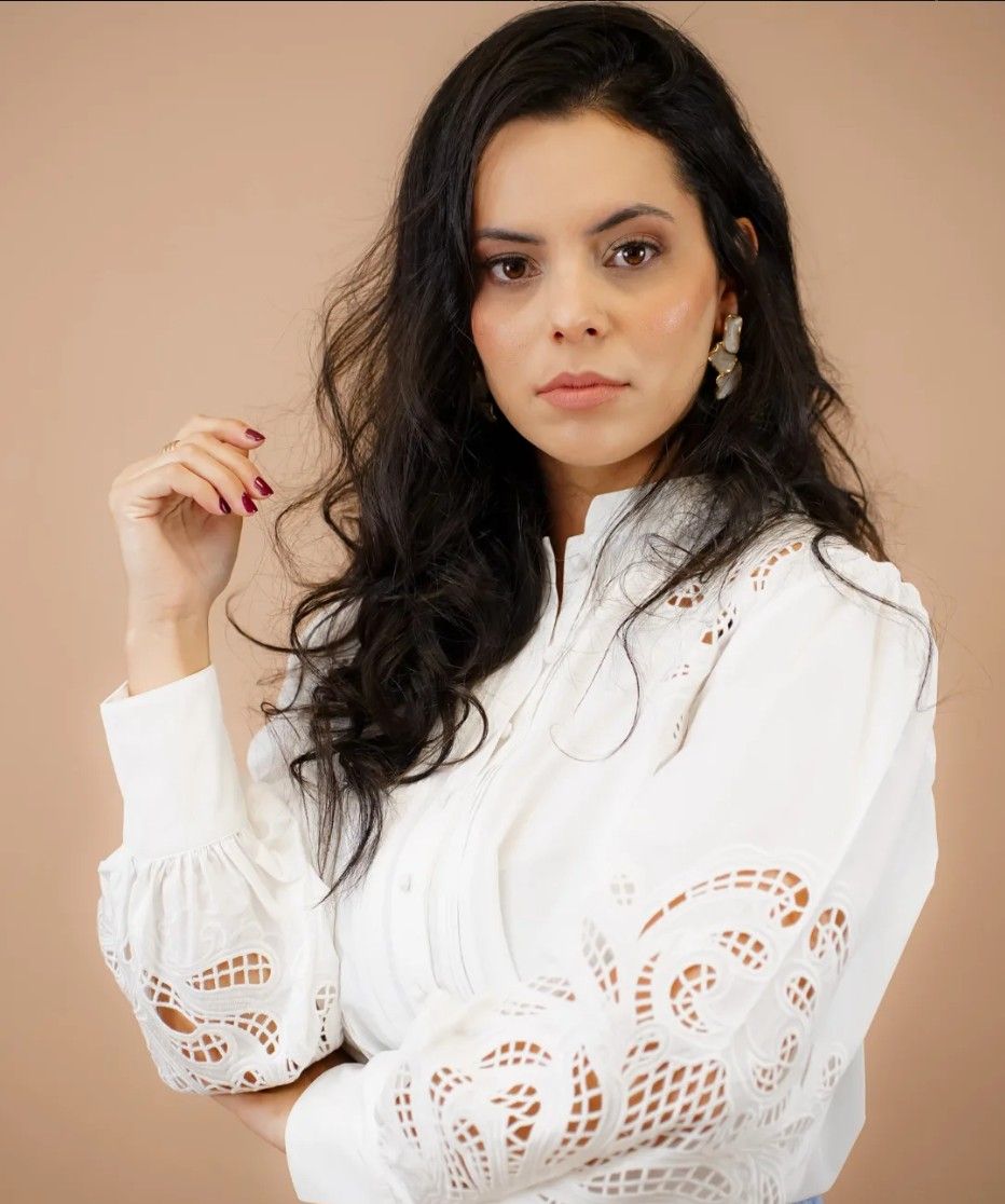 Retrato da fisioterapeuta Gabriela Alves, de cabelos longos escuros e blusa branca rendada, sobre fundo em tom nude