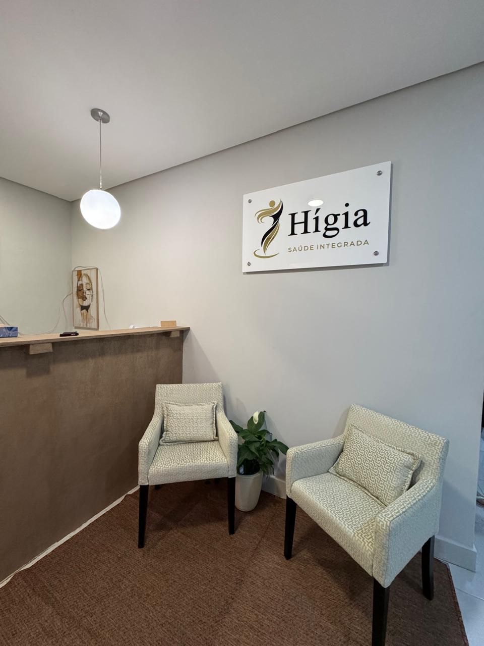 Recepção da Hígia com placa do logotipo na parede, duas poltronas claras e uma planta